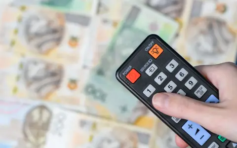 Dłoń trzyma pilota na tle polskich banknotów. Czy abonament RTV i zameldowanie mają związek z kosztami?