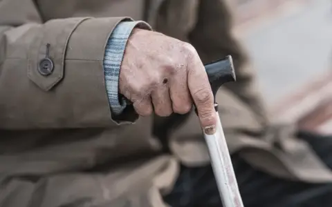 Dłoń starszego mężczyzny z widocznymi oznakami zespołu kruchości, mocno trzymająca laskę.