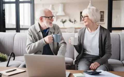 Para seniorów rozmawia przy kawie, planując pracę dla emeryta w domu. Na stole laptop i kalkulator.