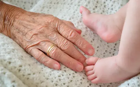 Dłoń starszej osoby z obrączką delikatnie dotyka stópki dziecka. Piękny obraz życia na emeryturze, pełnego miłości i nowych początków.