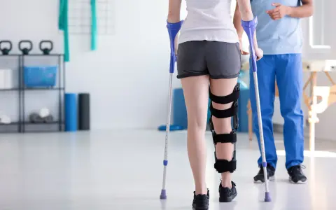 Fizjoterapeuta uczy, jak chodzić o kulach, wspierając pacjentkę z ortezą na nodze w sali rehabilitacyjnej.