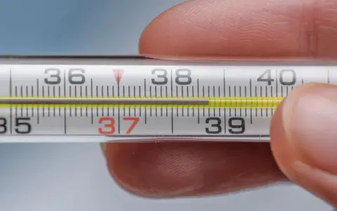 Zbliżenie na termometr rtęciowy, gdzie słupek rtęci wskazuje 37.1 temperatura. Dłoń trzyma termometr.