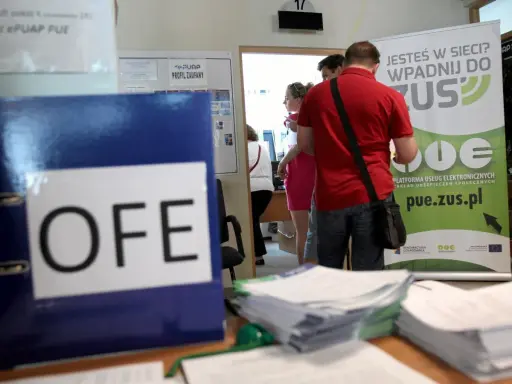 Na biurku leży segregator z napisem OFE. Ludzie stoją przy tablicy informacyjnej ZUS, pytając, czy można wypłacić pieniądze z OFE.
