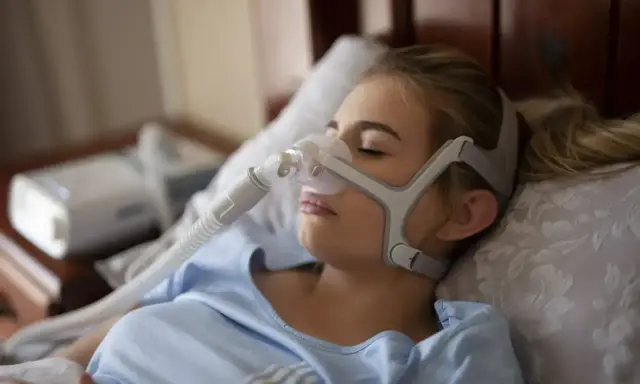Kobieta śpi w masce CPAP, która zapewnia wspomaganie oddychania w domu. Urządzenie pomaga jej spokojnie przesypiać noce.