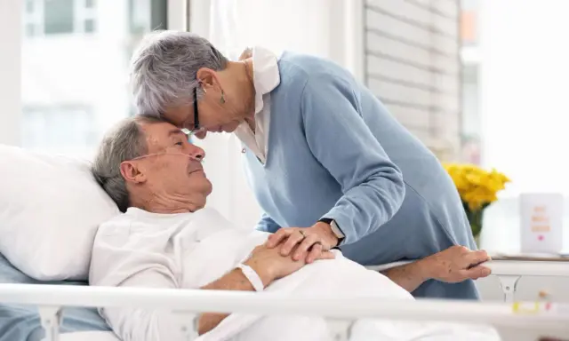 Czule pożegnanie. Kobieta pochyla się nad umierającym mężem, jak poznać że chory na alzheimera umiera, widać troskę i miłość.