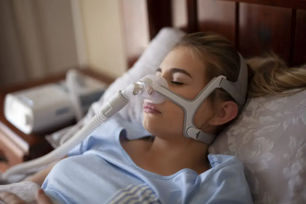 Młoda kobieta śpi w masce CPAP, kt&oacute;ra pomaga jej oddychać podczas snu, łagodząc bezdech nocny.