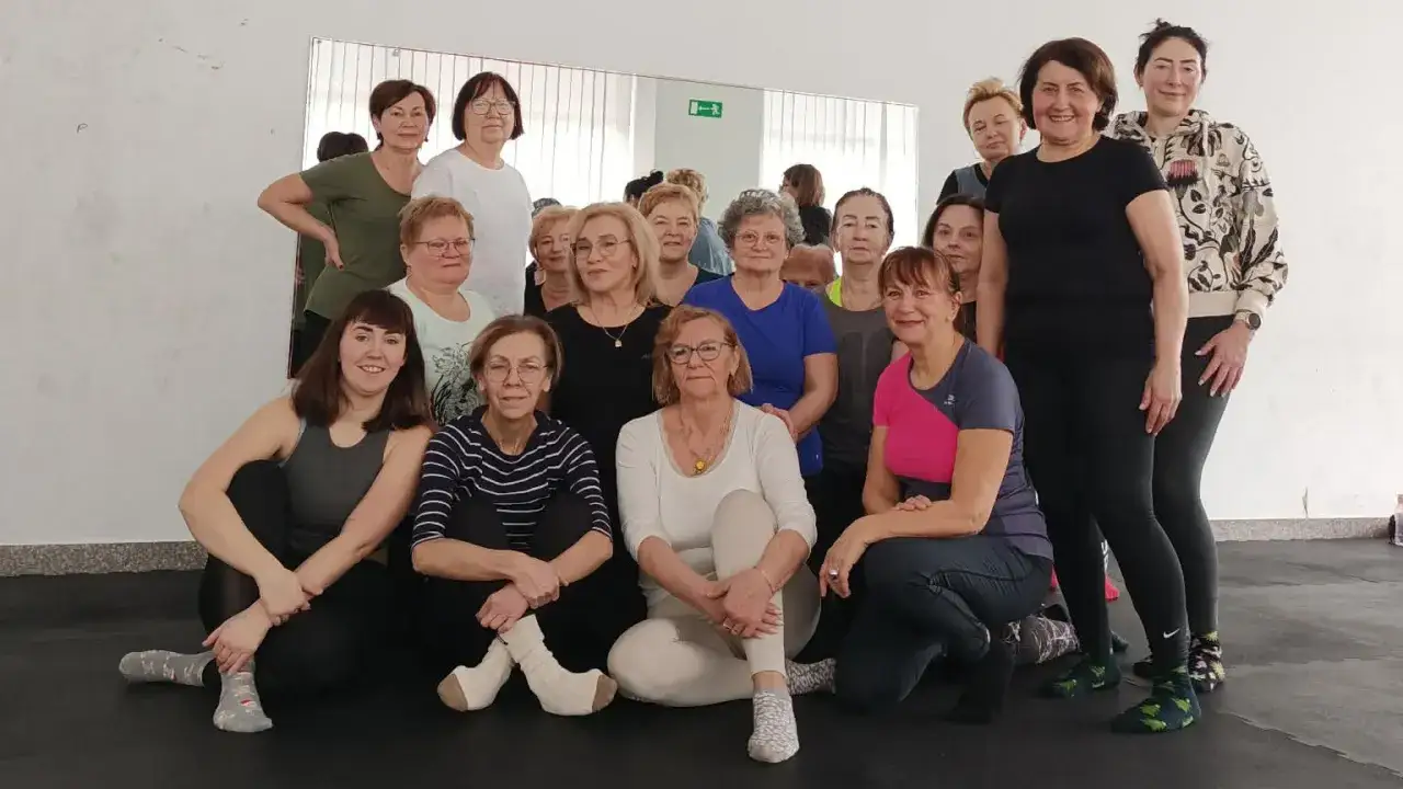 Grupa uśmiechniętych kobiet, uczestniczek zajęć dla senior&oacute;w, pozuje do zdjęcia po treningu.